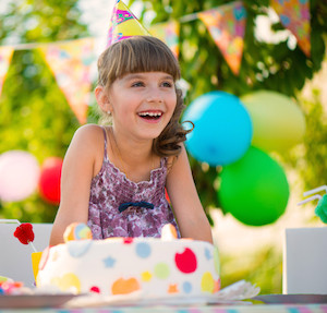 Little girl at a birthday party, laughing with a party hat and sitting in front of a cake with balloons in background